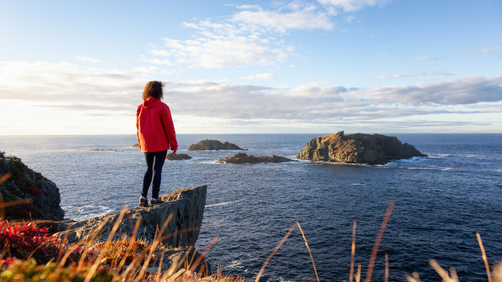Woman in red jacket overlooking rocky Newfoundland islands at sunset, showcasing coastal trails on Canada’s Atlantic Edge tours.