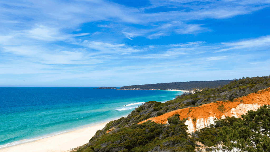 Scenic coastal cliffs and pristine beach on the Sapphire Coast Trek, showcasing vibrant red earth and blue ocean under clear skies.