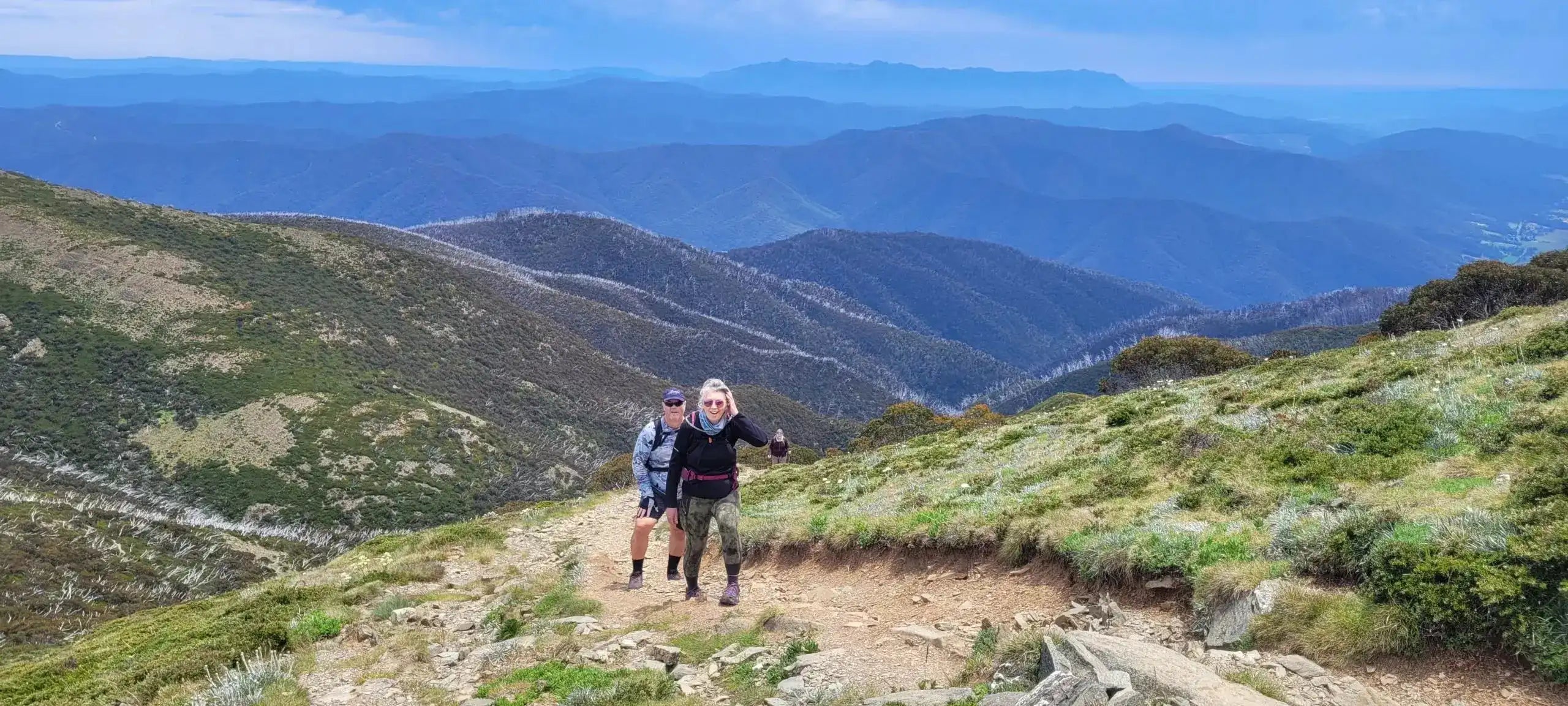 Two hikers ascending a trail with panoramic views of the Victorian Alps. Part of the moderate 4 Day High Country Horizons hiking adventure.