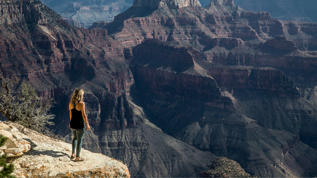 Woman stands on Grand Canyon cliff edge admiring vast red rock formations. Explore Western USA trips with canyon hikes and panoramic views.