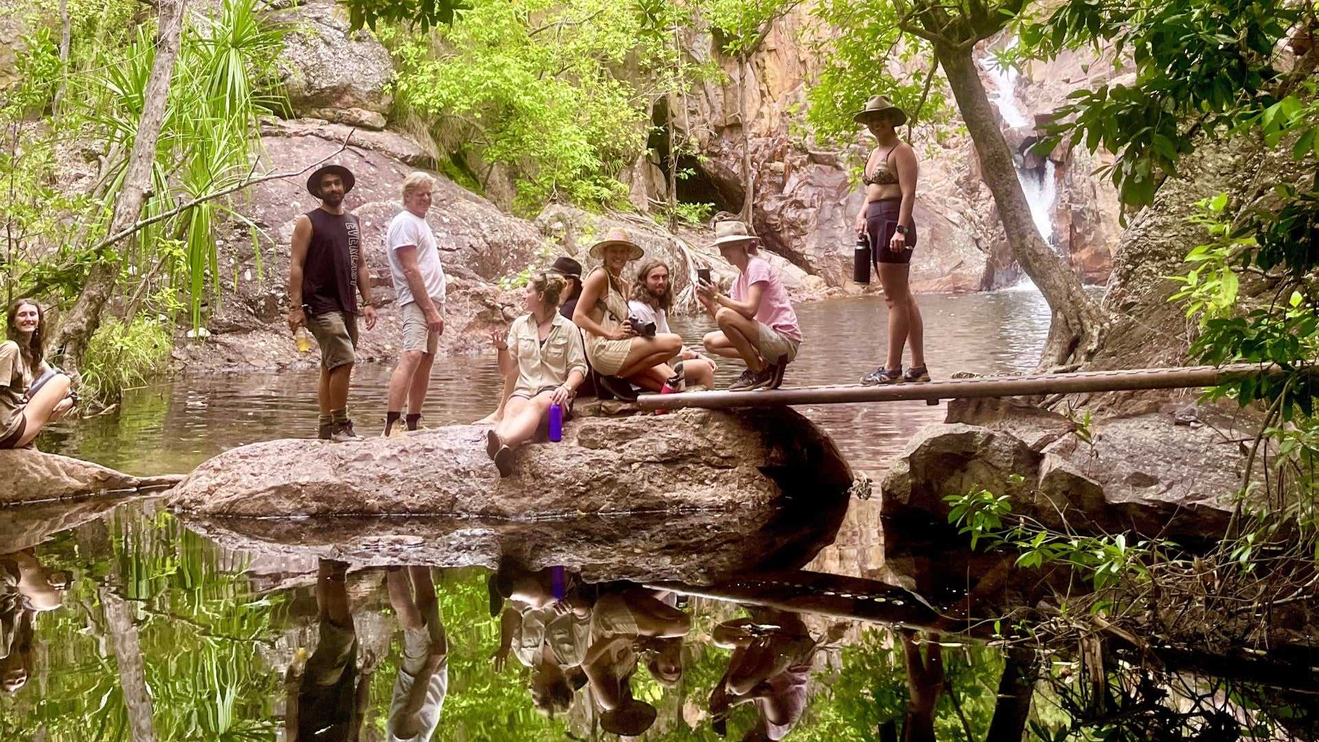 Tour group gathered by rock pool at Kakadu Litchfield Camping Adventure - 3-Day Tour. Natural setting with waterfall and forest backdrop.