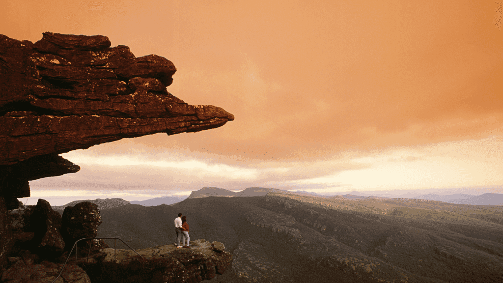 Couple standing on rock ledge overlooking stunning Grampians landscape during a colorful sunset.