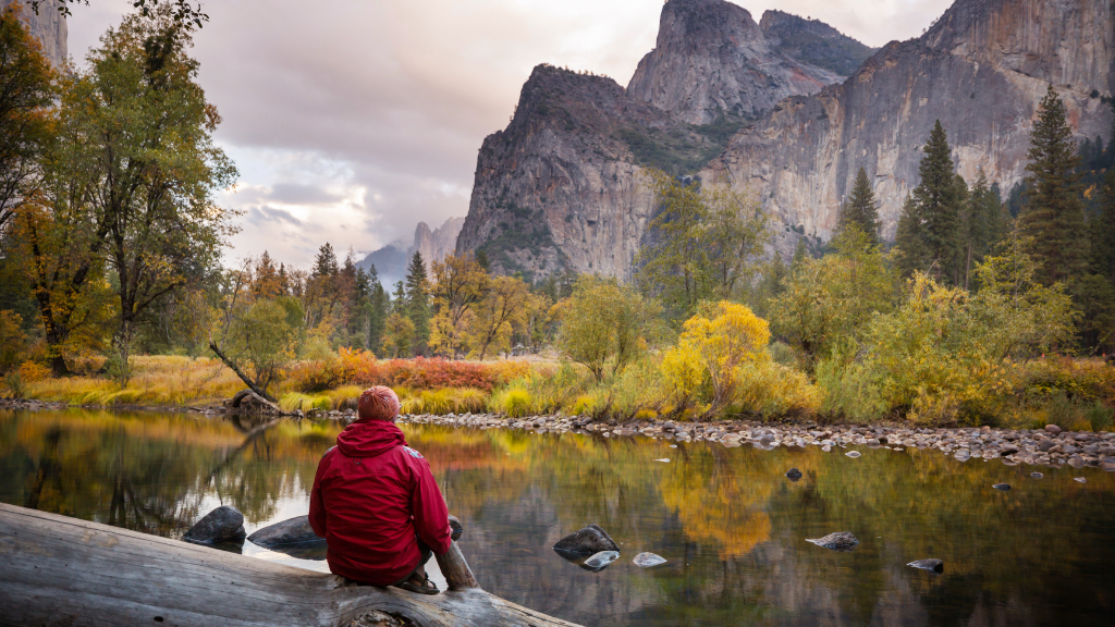 Traveler in red jacket sits by Yosemite Valley river at sunset, surrounded by fall foliage and granite cliffs. Explore Western USA trips with nature views.