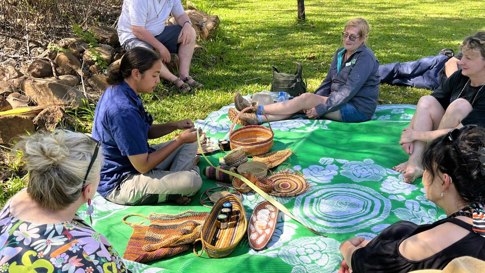 Tourists sit on picnic blanket learning Aboriginal weaving on Kakadu Katherine Gorge Tour | 4-Day Comfort Travel. Cultural workshop outdoors.