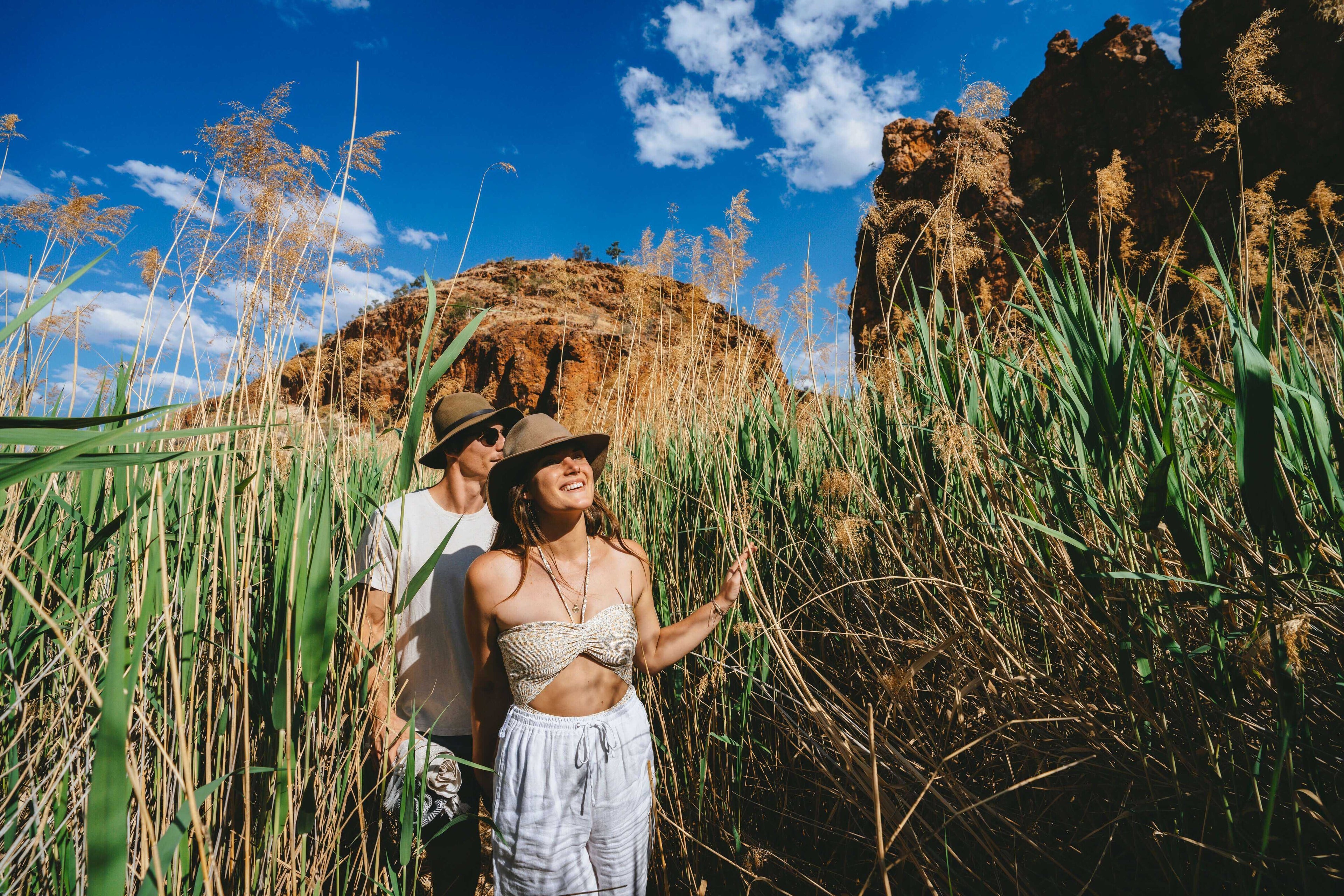 Couple walking through tall grass near red rock formations on Larapinta Trail 5-Day Expedition, ideal for nature lovers.