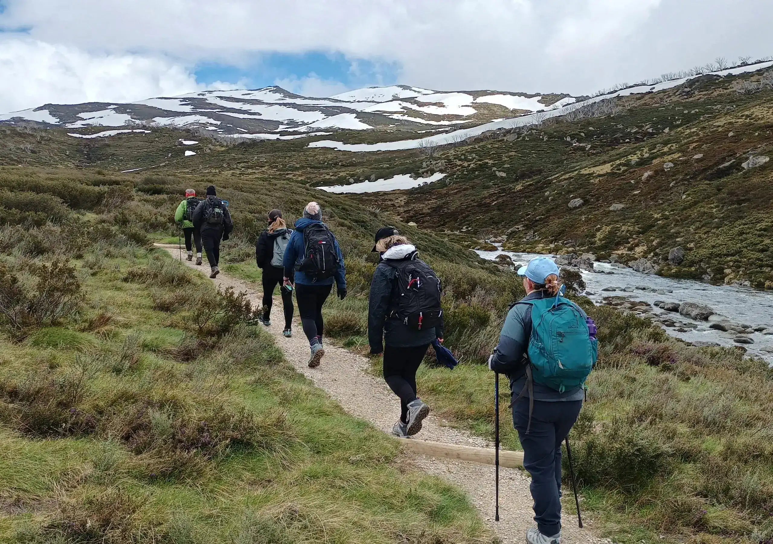 Group of hikers walking on a trail in a mountainous area with snow patches.