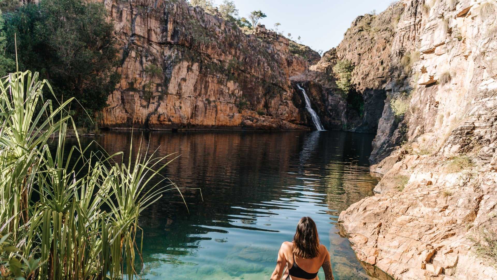 Woman entering clear plunge pool at Kakadu Litchfield Camping Adventure - 3-Day Tour. Tall waterfall and red rock cliffs in tranquil setting.