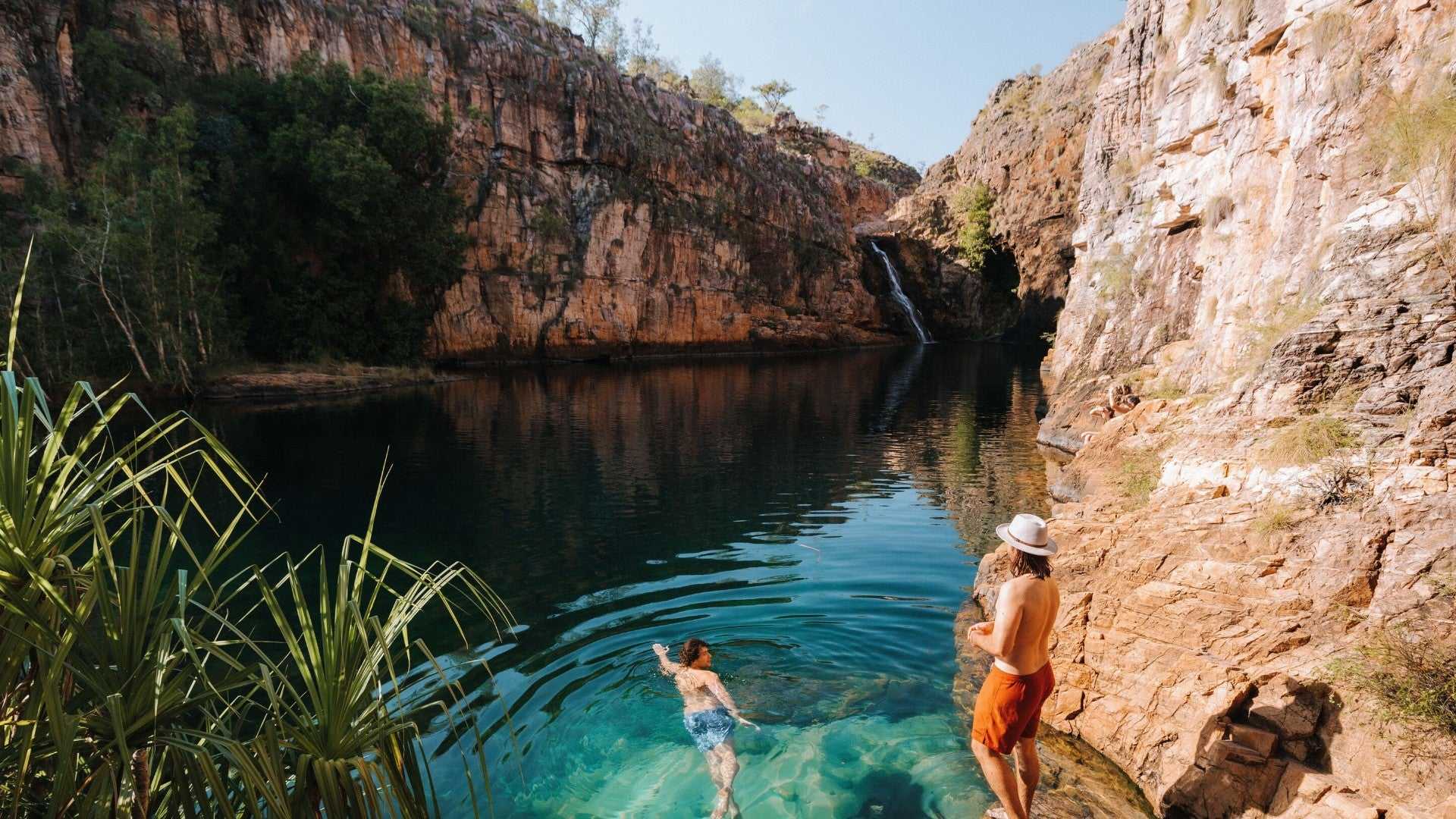 Man swimming in plunge pool beneath waterfall at Kakadu Litchfield Camping Adventure - 3-Day Tour. Scenic gorge with red cliffs and clear water.