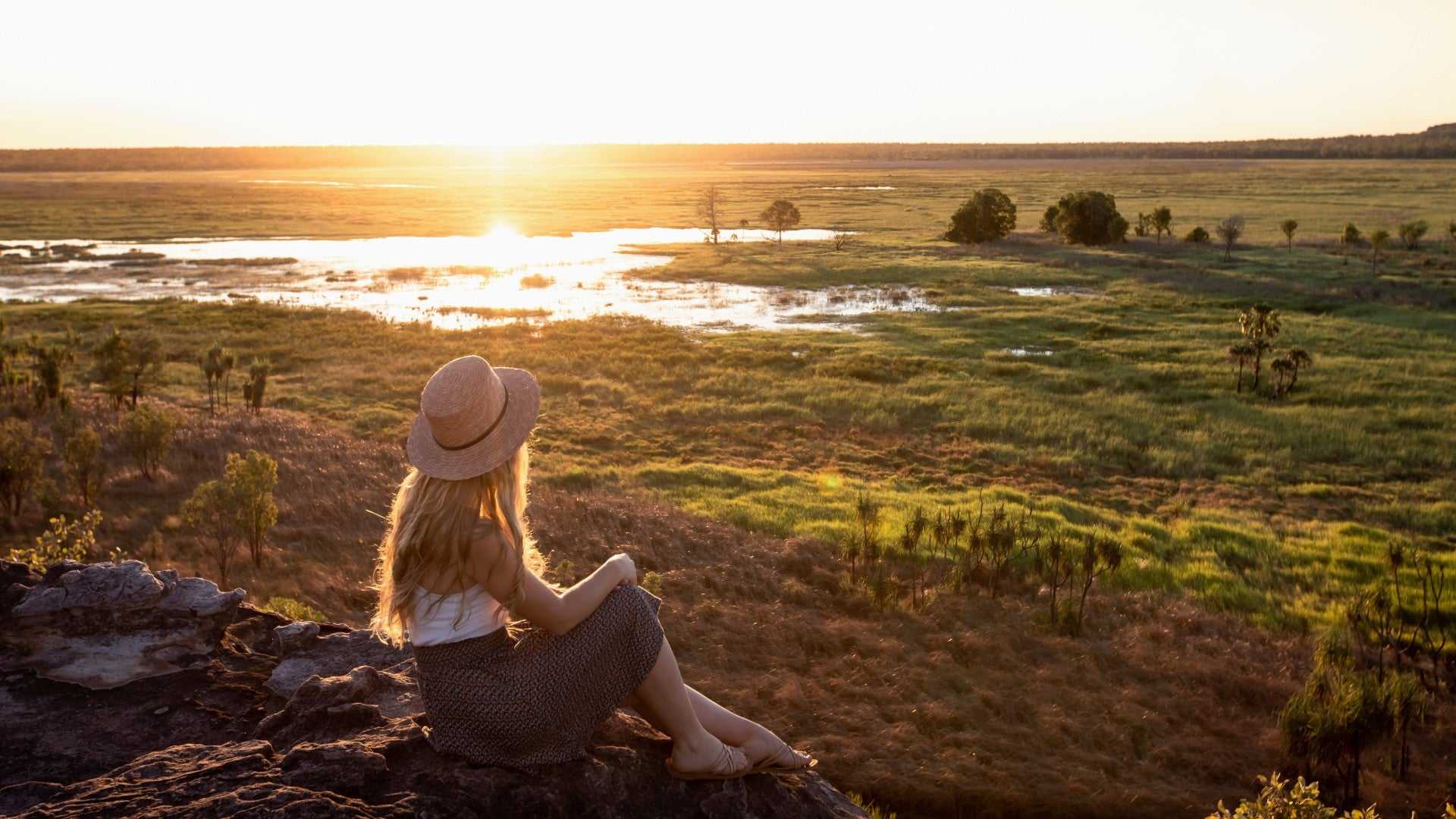 Woman watches sunset over wetlands from rocky ledge on Kakadu Katherine Gorge Tour | 4-Day Comfort Travel. Peaceful end to adventure day.