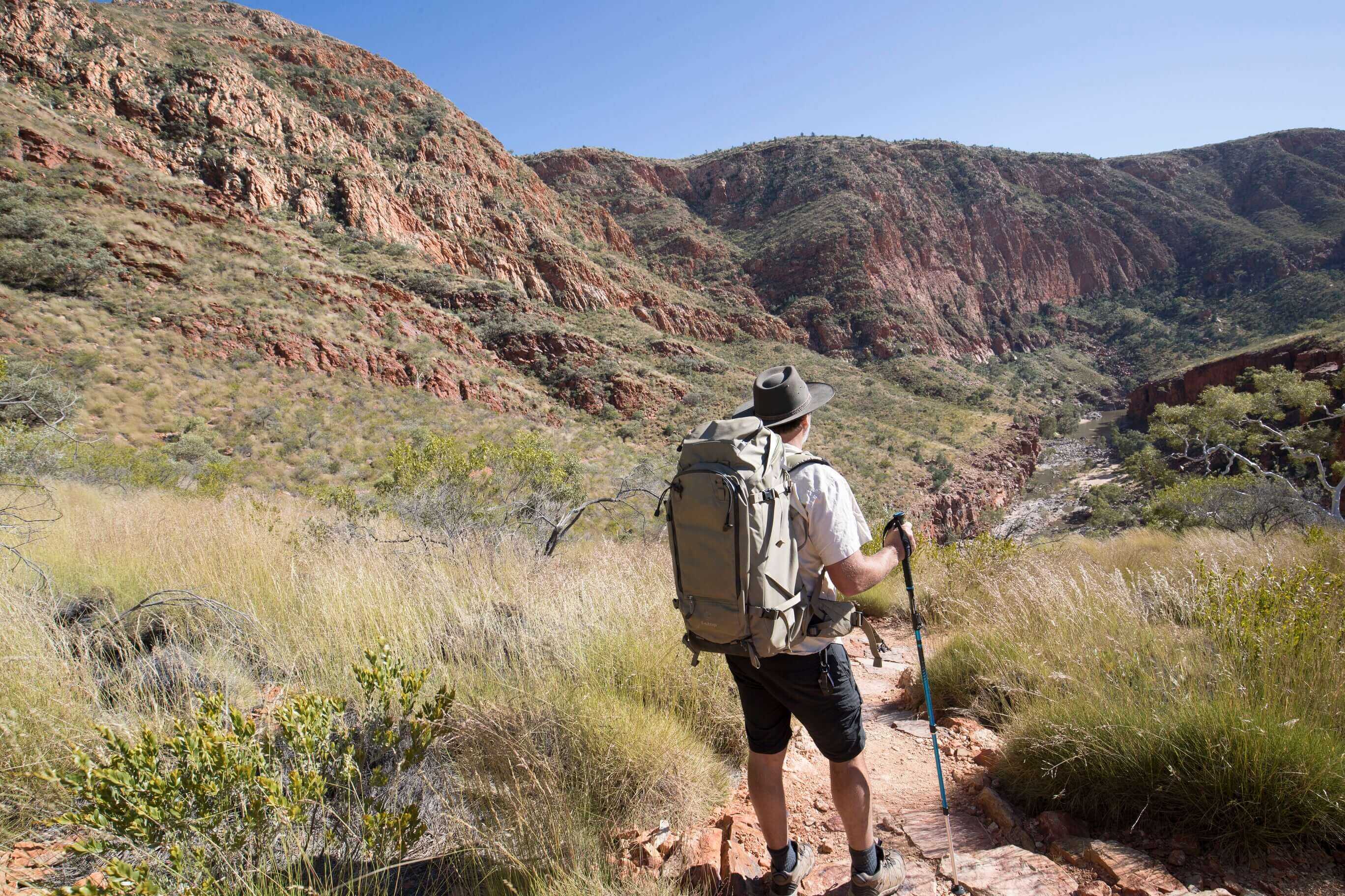 Solo hiker with backpack walking through red rock valley, embracing solitude on Larapinta Trail 5-Day Expedition in Australia.