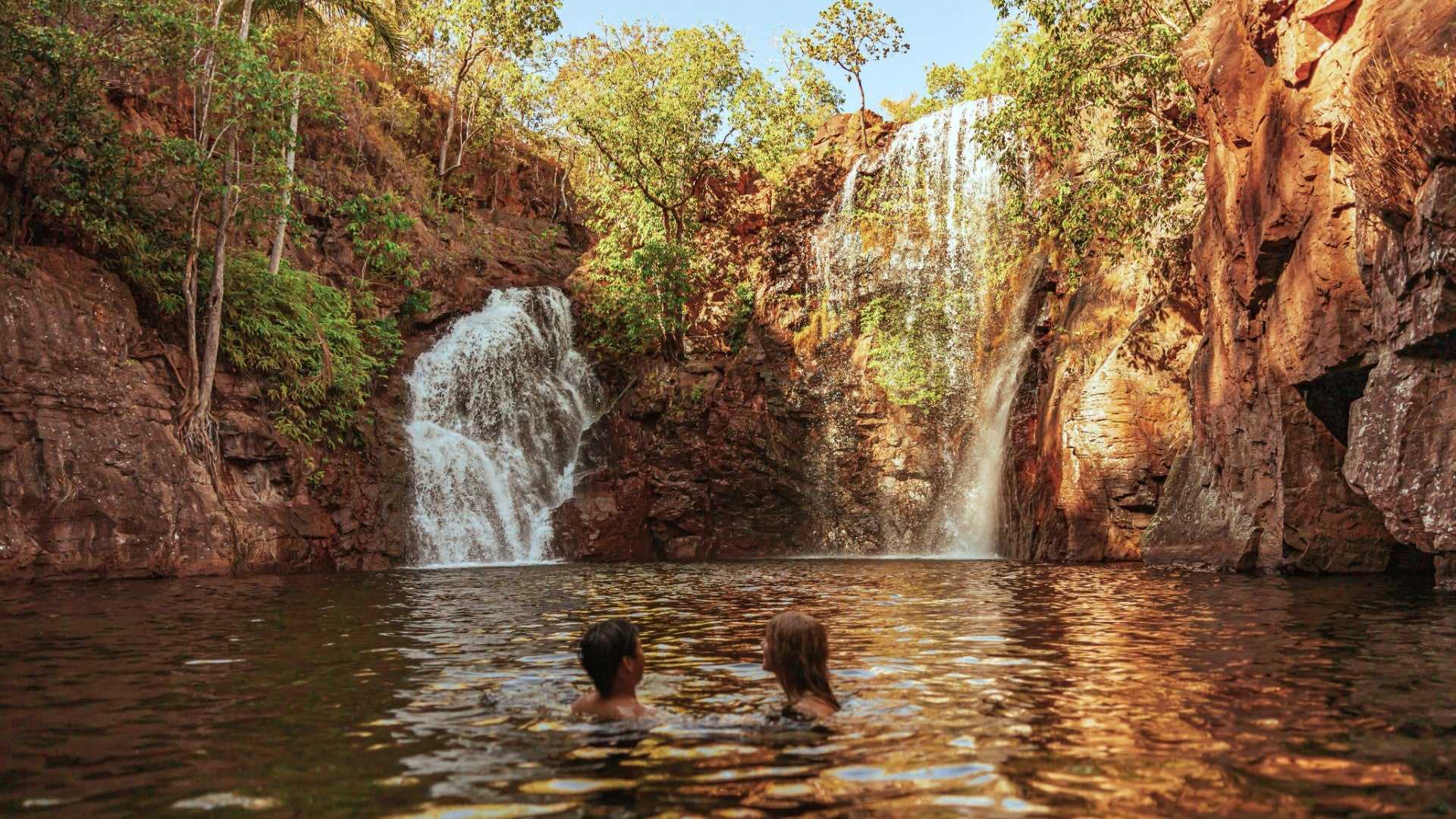 Couple swimming near twin waterfalls at Kakadu Litchfield Camping Adventure - 3-Day Tour. Refreshing natural pool surrounded by red rock walls.