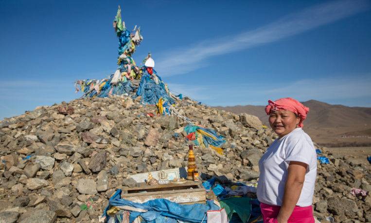 Local woman standing beside a sacred ovoo stone shrine in Mongolia, part of the cultural highlights on the Steppe to Sand - Mongolia Gobi Desert Tour.