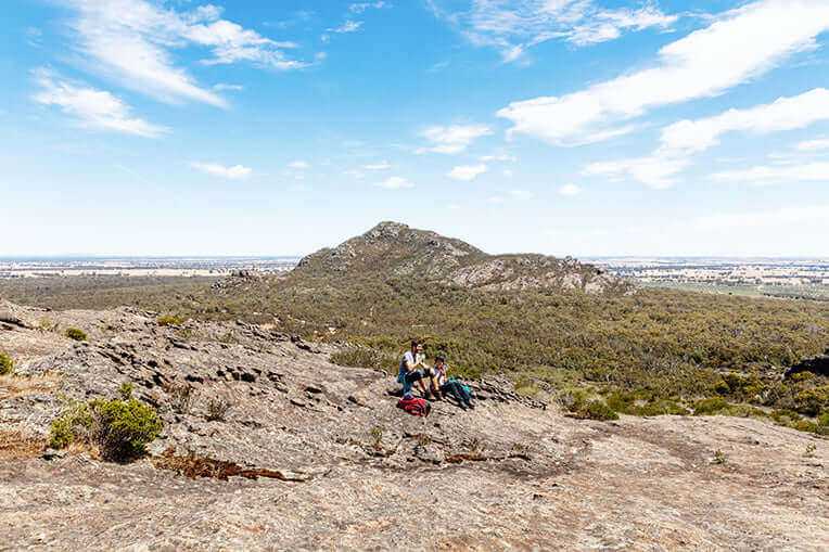 Adventure seekers relaxing on rocky summit in Grampians, enjoying stunning views during guided outdoor tour.