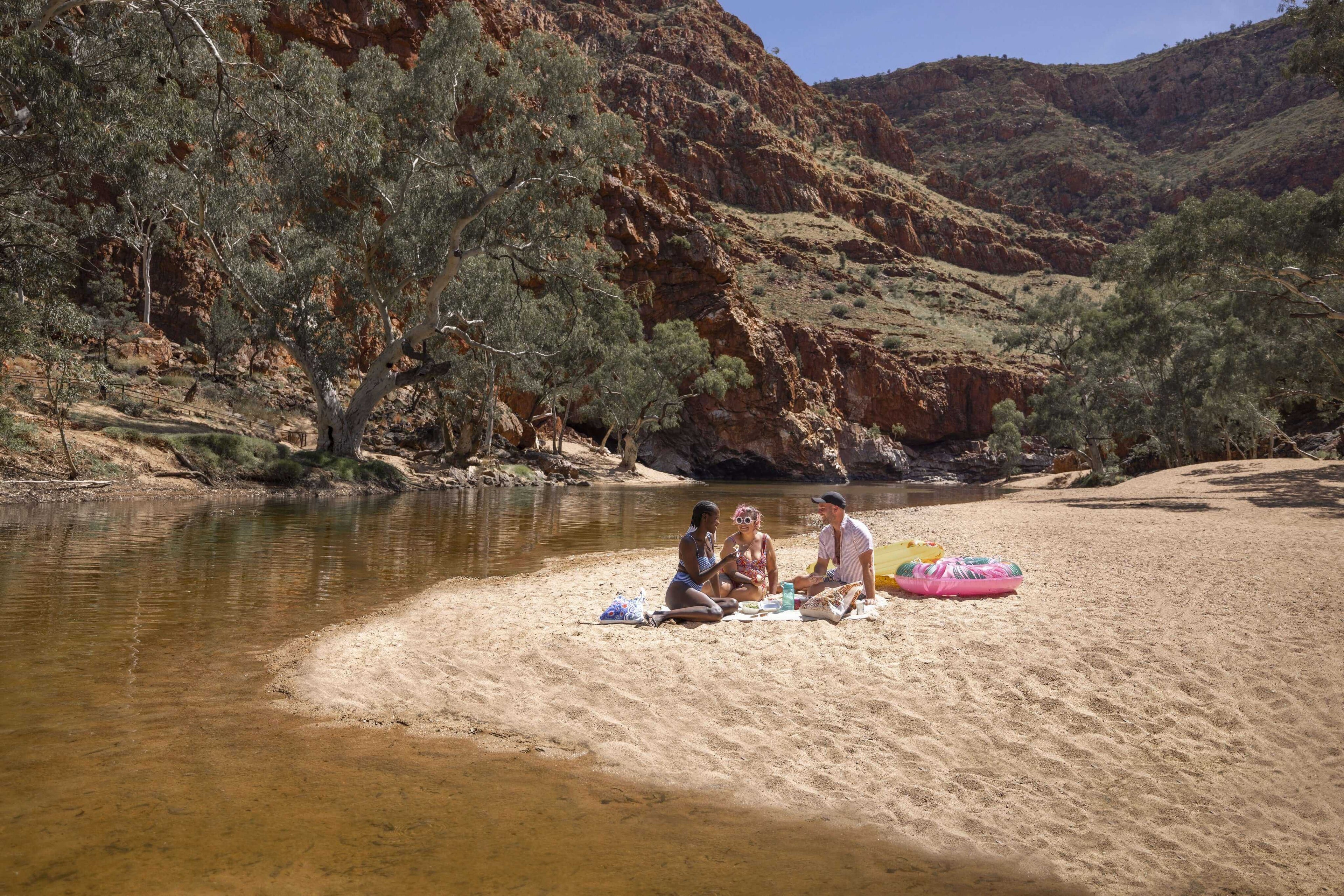 Family enjoying picnic by sandy riverbank in West MacDonnell Ranges, a relaxing stop on Larapinta Trail 5-Day Expedition.