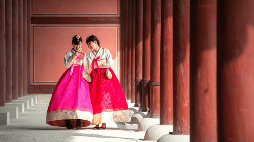 Two women in vibrant hanbok walk through a traditional palace corridor in Seoul on a South Korea cultural tour. Experience ancient Korean heritage.