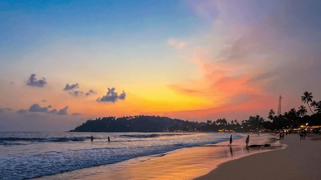 Sunset over a tropical beach with people in the surf, a relaxing end to the 12-day Sri Lanka spice gardens and coast tour.