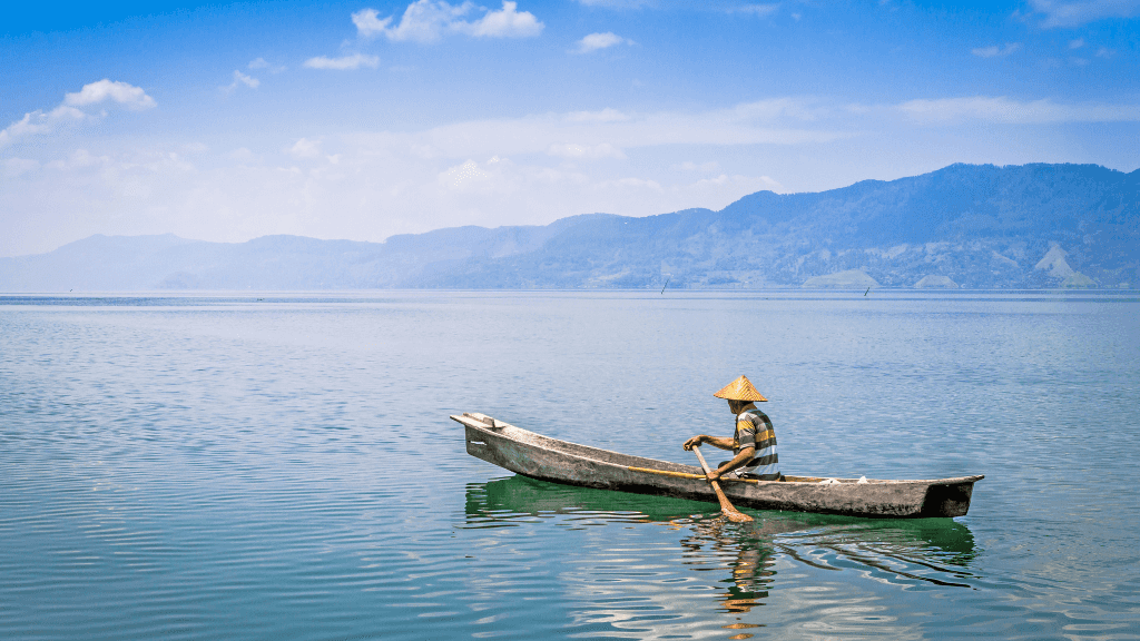Local fisherman paddling wooden canoe on calm lake with mountain backdrop, part of the Sumatran Jungle River Expedition tour.