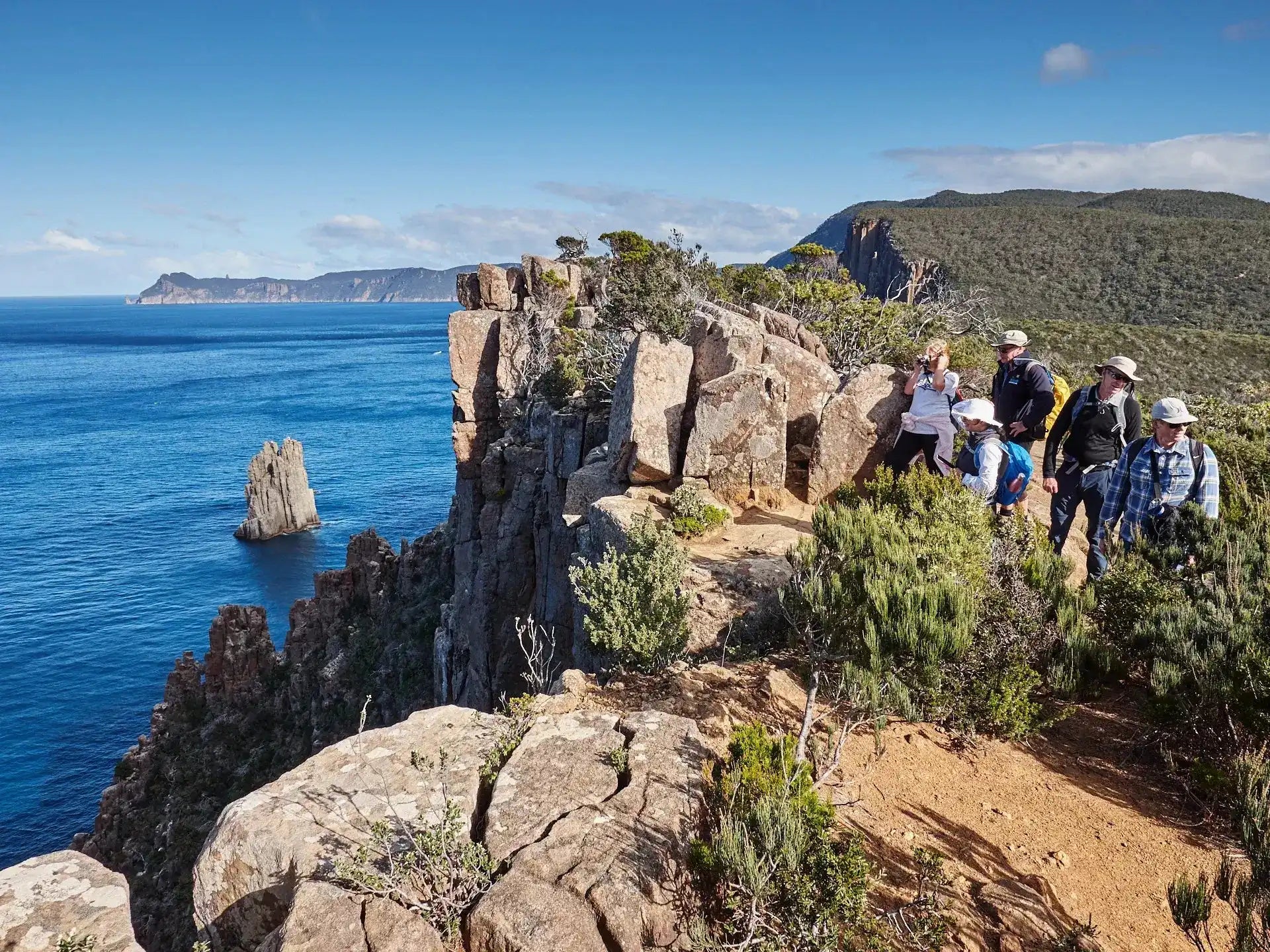 Group of people hiking on a rocky cliff overlooking the ocean with mountains in the background.