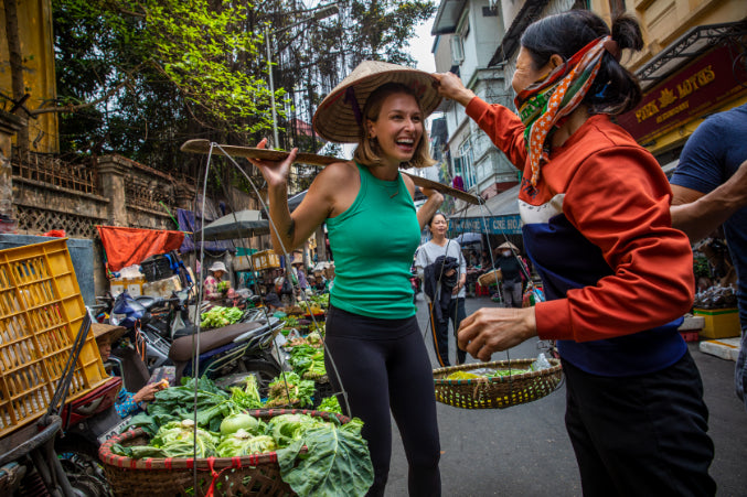 Local vendor placing a conical hat on a smiling tourist. Authentic market visit on Vietnam Hiking and Cycling Tour.