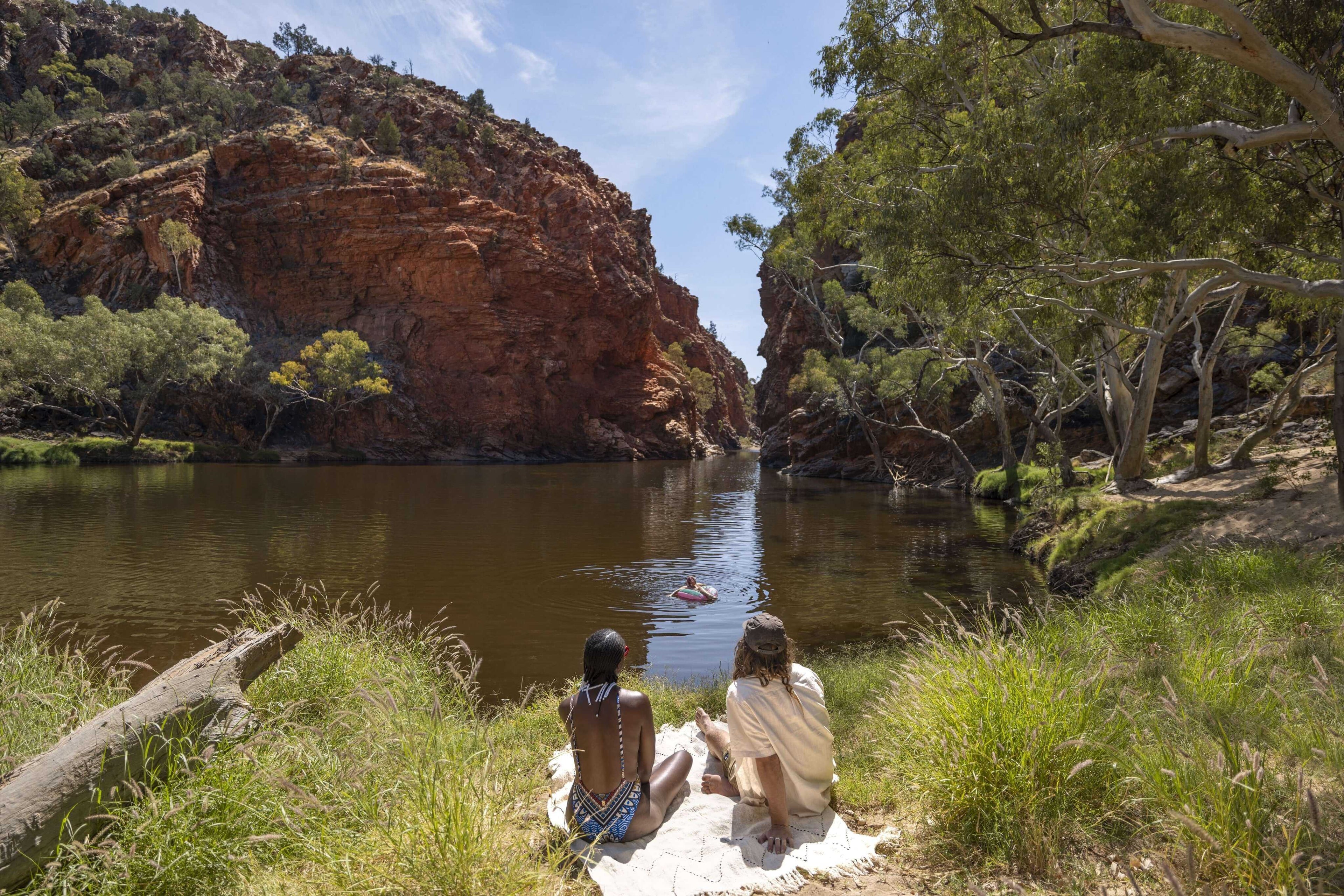 Two people relaxing by waterhole surrounded by cliffs, a peaceful moment on Larapinta Trail 5-Day Expedition in Australia.