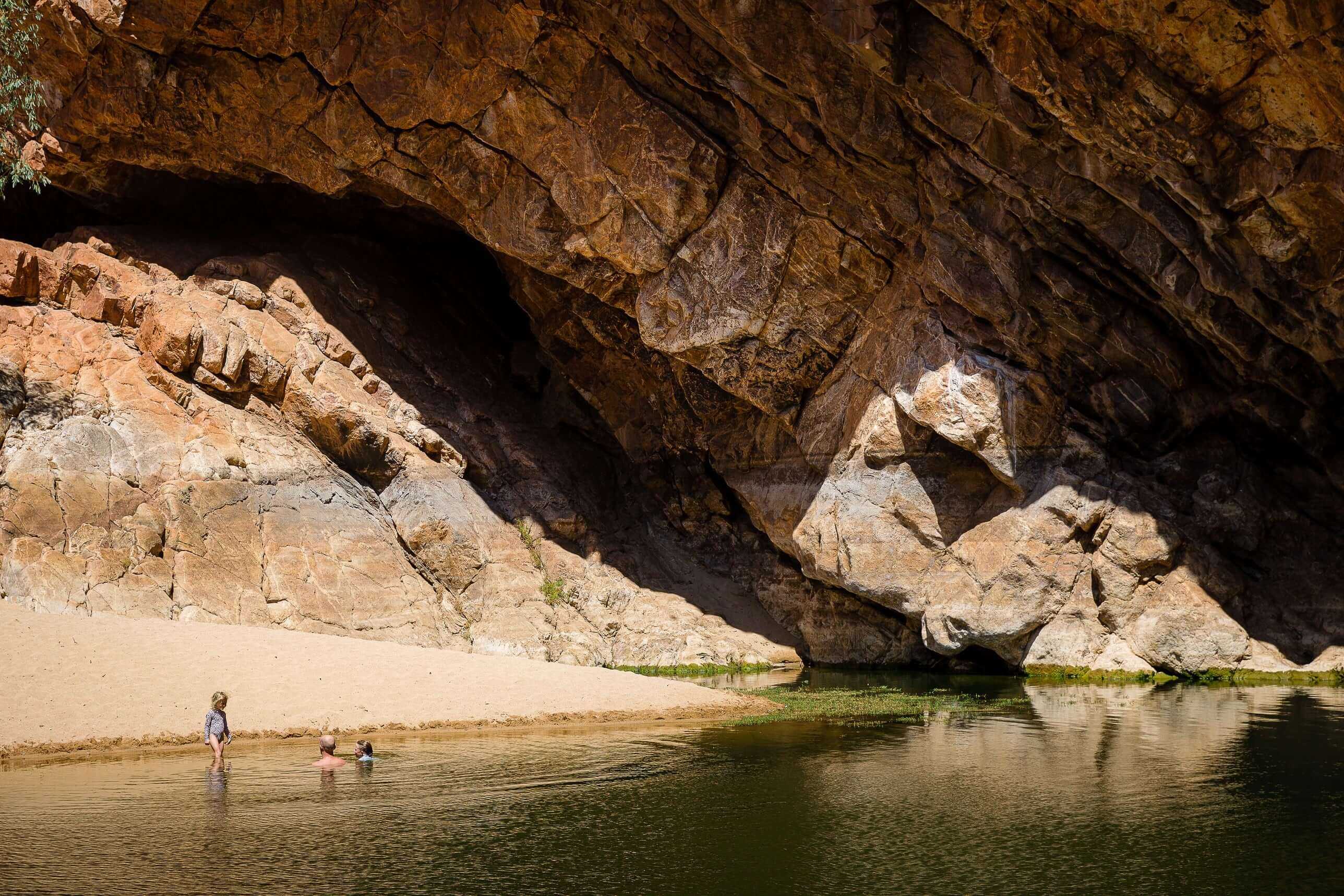 Children playing in waterhole under rock overhang, a family-friendly stop on Larapinta Trail 5-Day Expedition in Australia.