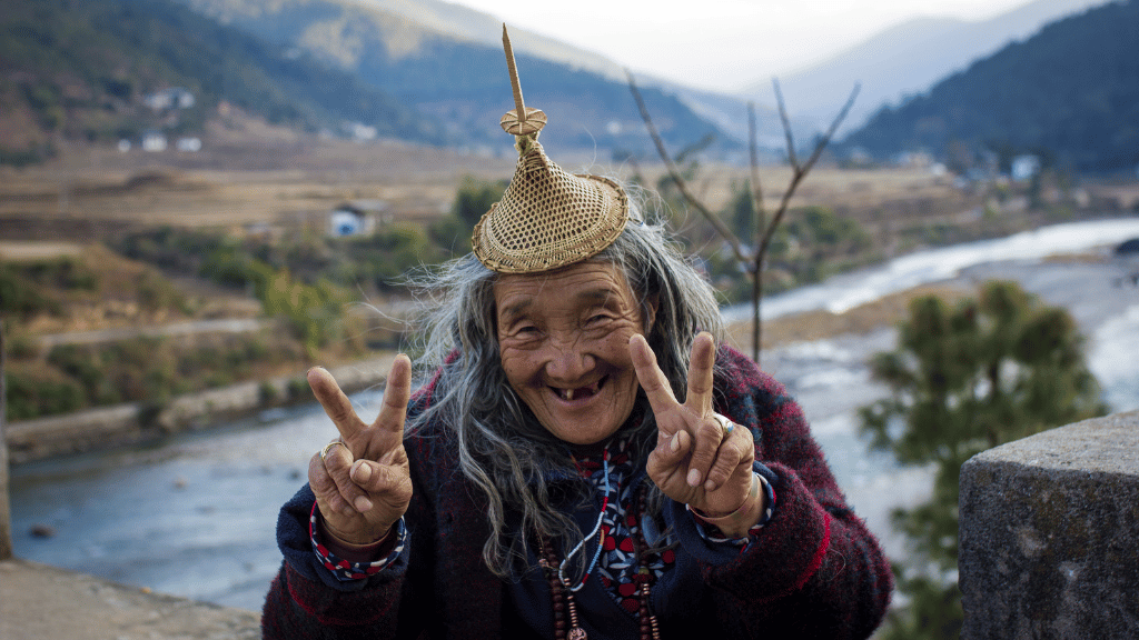 Elderly Bhutanese woman smiling and flashing peace signs, reflecting the warmth of local culture on the trail.
