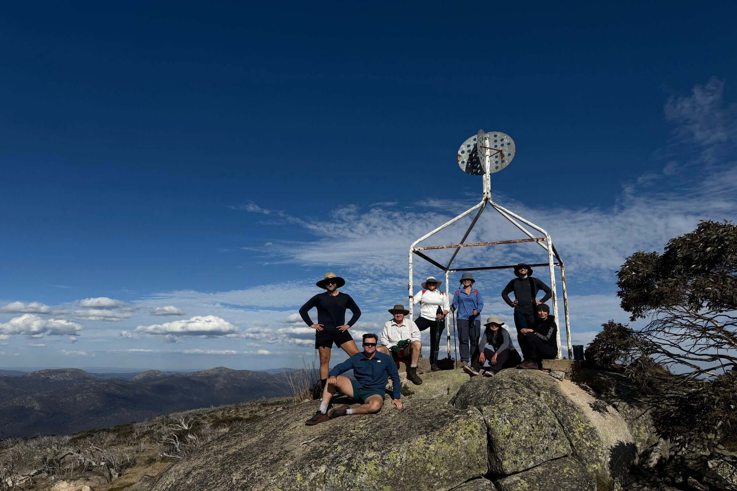 Group posing at Mt Bimberi summit trig station with scenic mountain views on guided outdoor adventure tour.