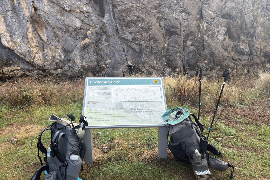 Coolaman Cave sign with hiking gear in front of limestone cliffs on Mt Bimberi & Blue Waterholes tour route.