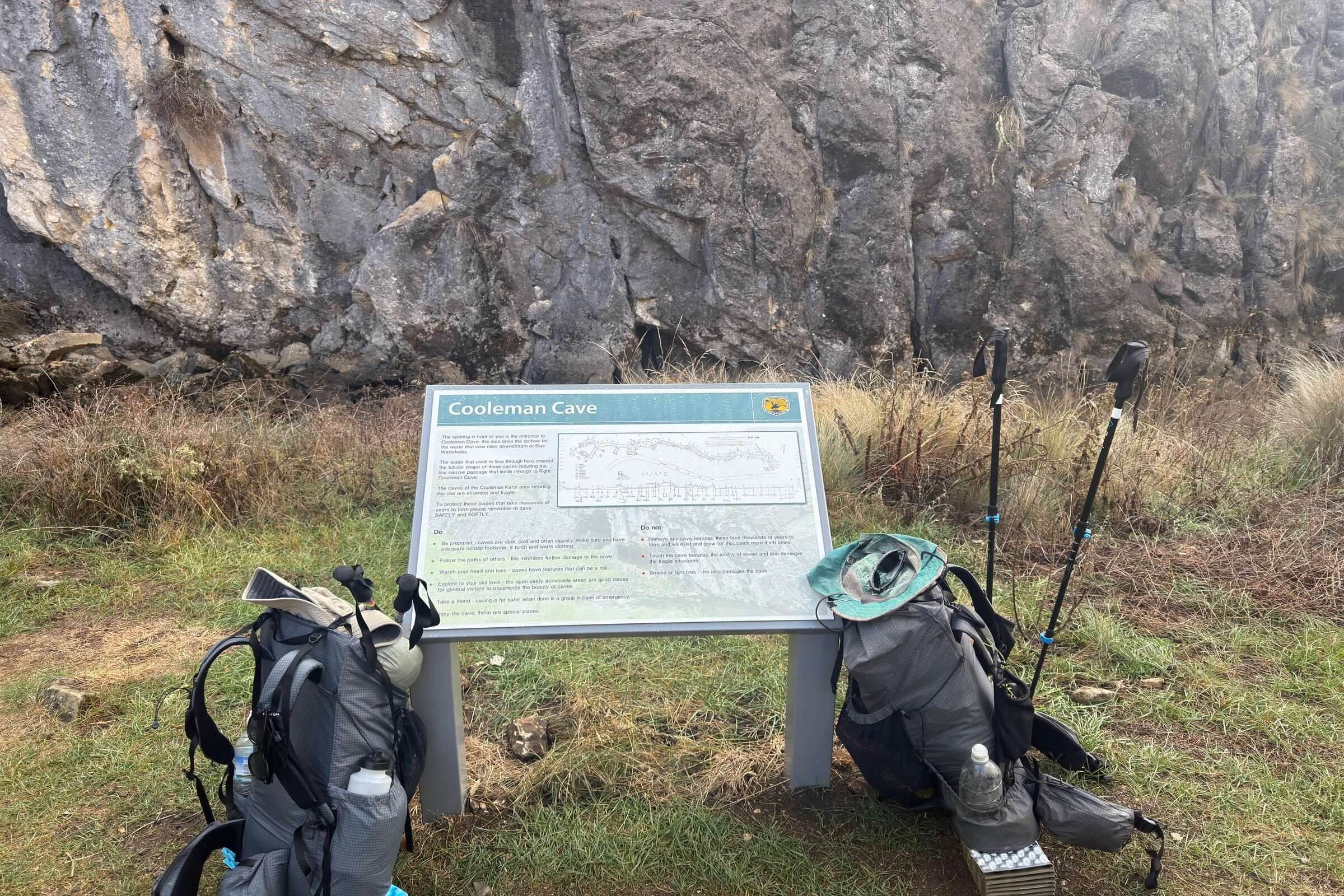 Coolaman Cave sign with hiking gear in front of limestone cliffs on Mt Bimberi & Blue Waterholes tour route.