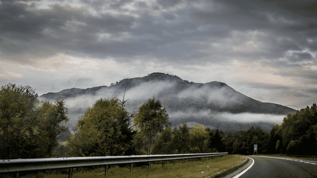 Misty mountain view alongside a winding road, surrounded by lush trees and a dramatic sky, perfect for outdoor adventures.