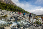 Group of hikers on a wooden bridge over a stream with mountains in the background