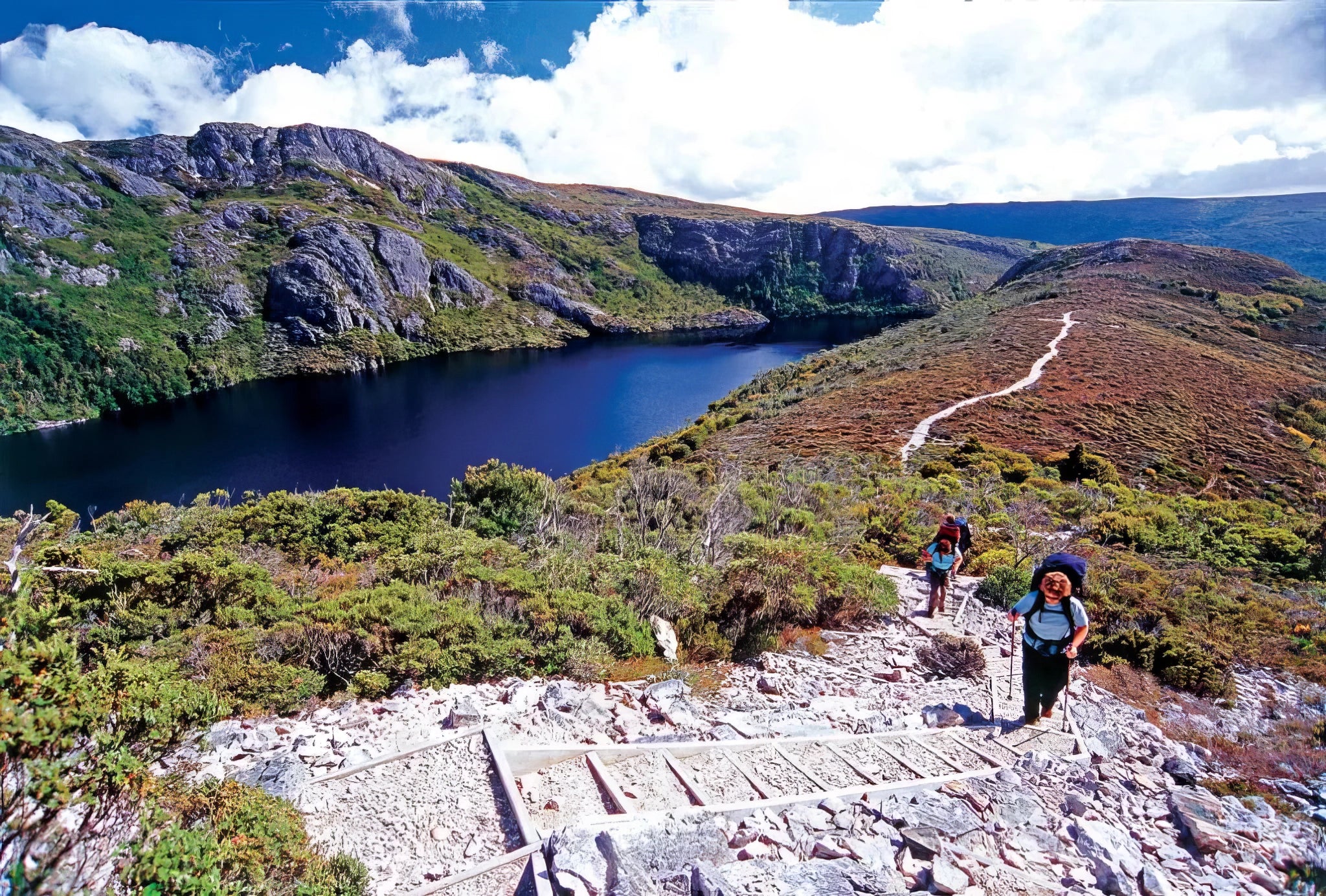 Two hikers on a trail near a lake surrounded by mountains
