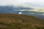 Two hikers on a grassy hill with a scenic view of a river and forest.