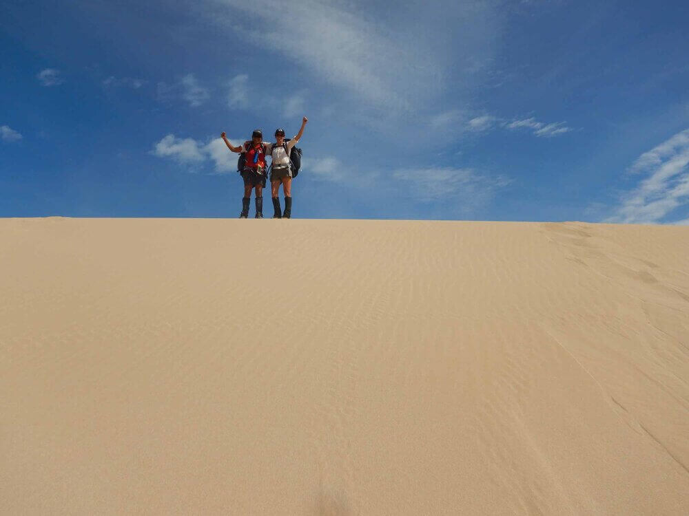 Two hikers celebrating atop a sand dune under a blue sky during an adventurous outdoor trek.