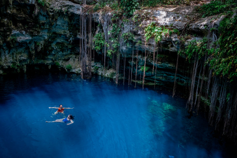 Two people swimming in a deep blue cenote with rocky cliffs and vegetation.