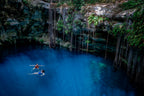 Two people swimming in a deep blue cenote with rocky cliffs and vegetation.