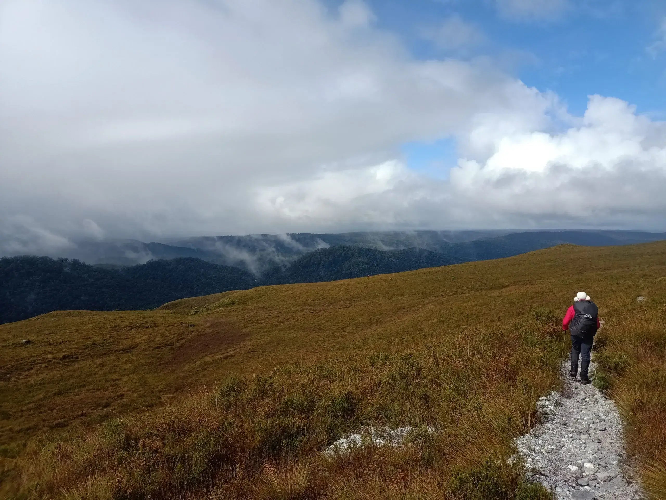 Person hiking on a trail in a mountainous landscape with clouds and blue sky.