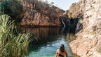 Woman entering clear plunge pool at Kakadu Litchfield Camping Adventure - 3-Day Tour. Tall waterfall and red rock cliffs in tranquil setting.