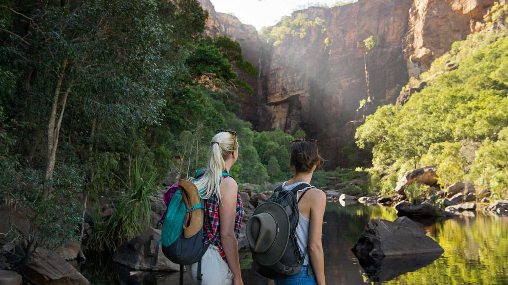 Two hikers admiring waterfall view at Kakadu Litchfield Camping Adventure - 3-Day Tour. Scenic bushwalk with towering cliffs and water.