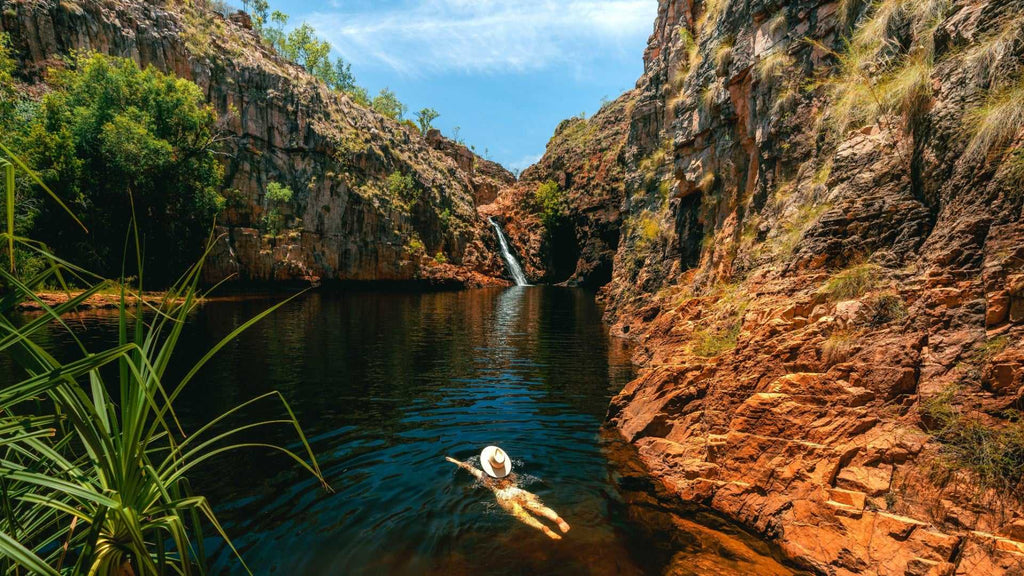 Traveler floating in plunge pool beneath waterfall at Kakadu Litchfield Camping Adventure - 3-Day Tour. Peaceful gorge with red rock walls.