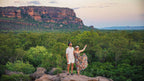 Couple admiring panoramic escarpment views during Kakadu Litchfield Camping Adventure - 3-Day Tour. Vast green forest and sandstone cliffs at sunset.