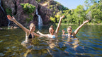Three women swimming in waterfall pool at Kakadu Litchfield Camping Adventure - 3-Day Tour. Lush green backdrop and joyful moment.