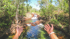 Hands framing cascading stream at Kakadu Litchfield Camping Adventure - 3-Day Tour. Clear water flows through rocky forest terrain.