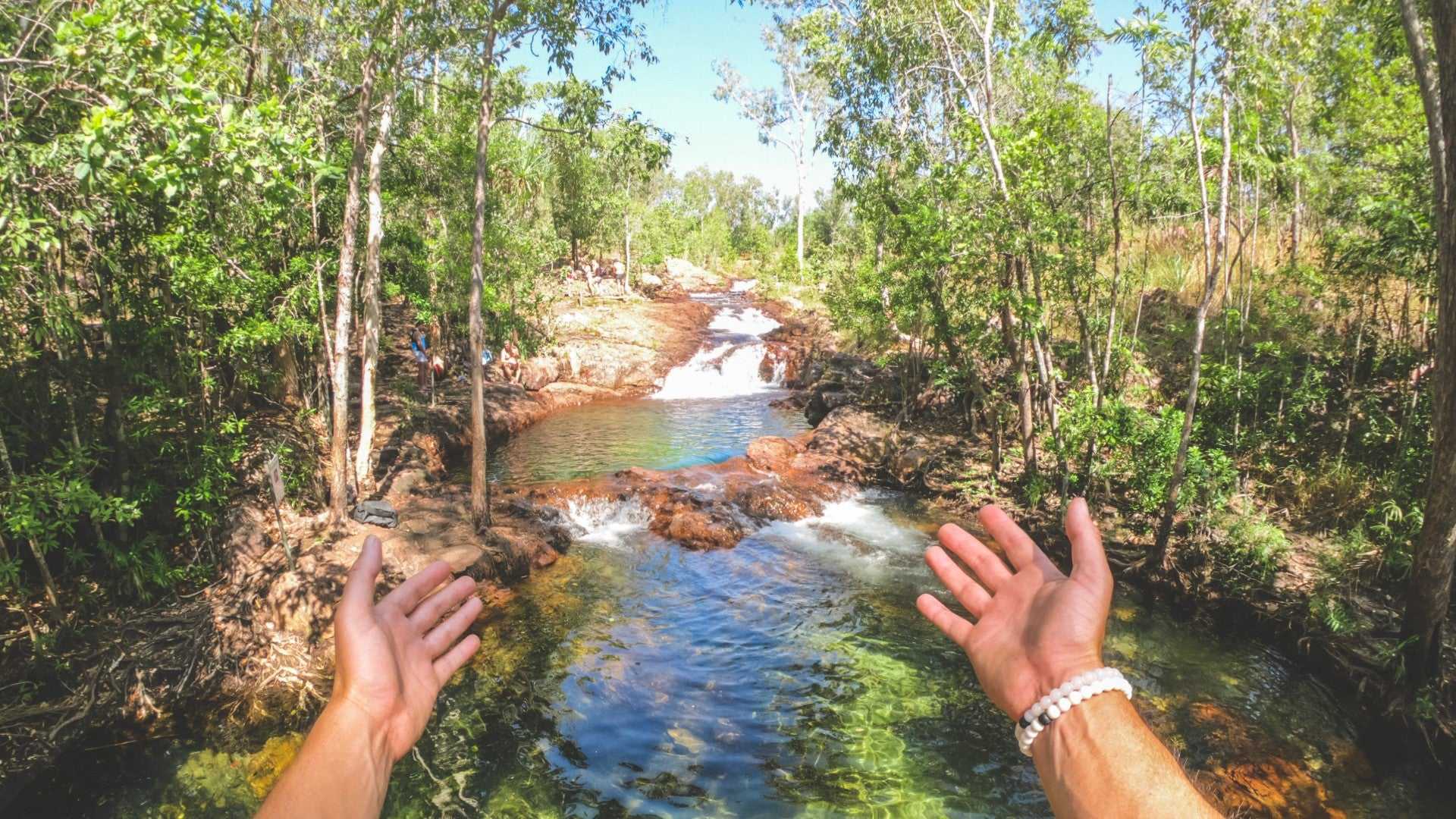 Hands framing cascading stream at Kakadu Litchfield Camping Adventure - 3-Day Tour. Clear water flows through rocky forest terrain.