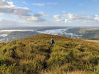 Person standing on a grassy hill with a scenic view of valleys and clouds.