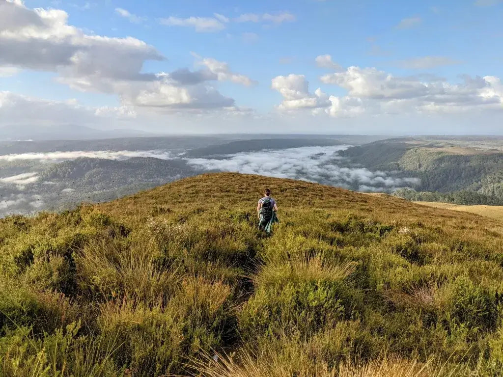 Person standing on a grassy hill with a scenic view of valleys and clouds.