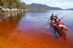 Two people in a kayak on a calm water body with mountains in the background