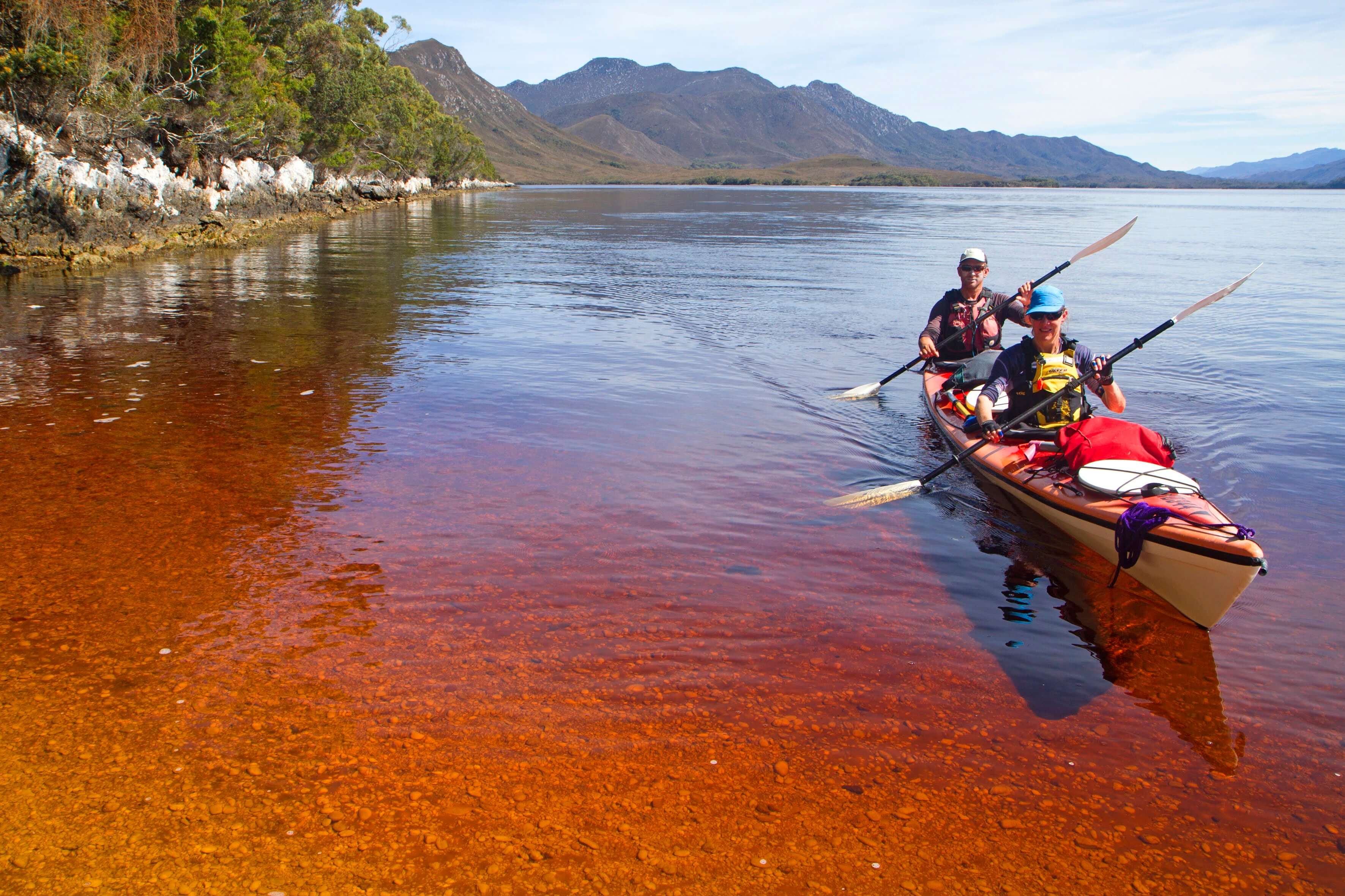 Two people in a kayak on a calm water body with mountains in the background