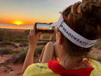 Woman capturing a sunset with her phone, wearing a Headsweats visor in the great outdoors.