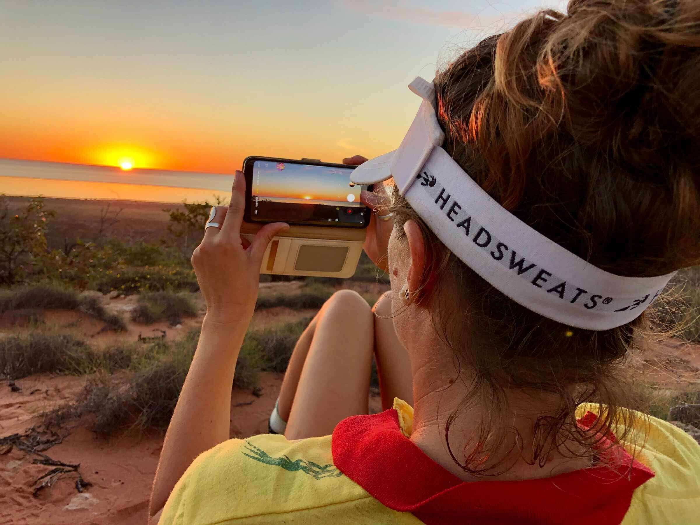Woman capturing a sunset with her phone, wearing a Headsweats visor in the great outdoors.
